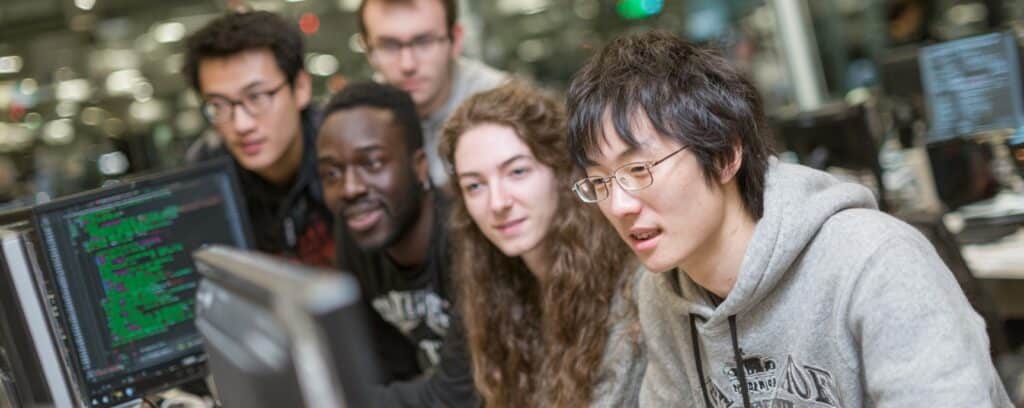 Five students sit closely together, looking at computer monitors with code on the screens in a tech lab.