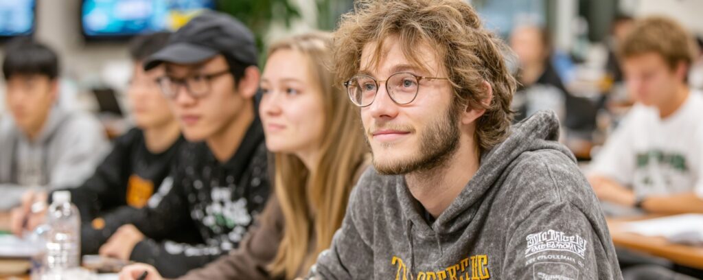 Students sitting in a classroom, attentively listening, with one student in focus wearing glasses and a hoodie.